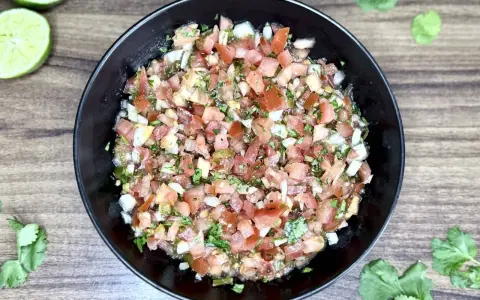 Świeże, kolorowe pico de gallo w czarnej misce, z limonkami i kolendrą na drewnianym tle.