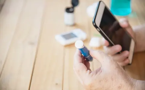 Osoba z cukrzycą sprawdza poziom glukozy, trzymając telefon. W tle glukometr i inne akcesoria. Czy alkohol w wydychanym powietrzu wpływa na wyniki?