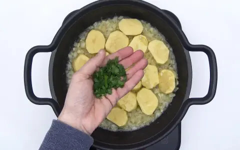 Manos añaden perejil picado a patatas en rodajas sobre cebolla sofrita en una sartén. ¡El inicio de unas deliciosas patatas en salsa verde!