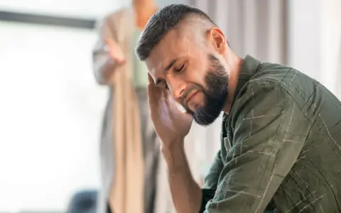 Mężczyzna z ręką na skroni, wyrażający zaangażowanie emocjonalne. W tle niewyraźna postać.