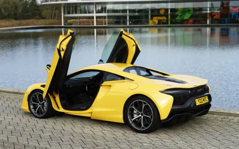 Żółty McLaren Artura z otwartymi drzwiami, stojący nad jeziorem przy nowoczesnym budynku.