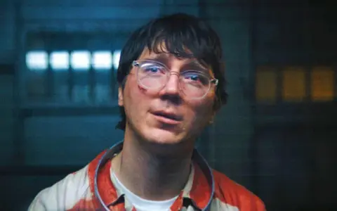 Paul Dano med glasögon och blodfläckar på sin randiga skjorta, i en mörk miljö med ljus bakom.