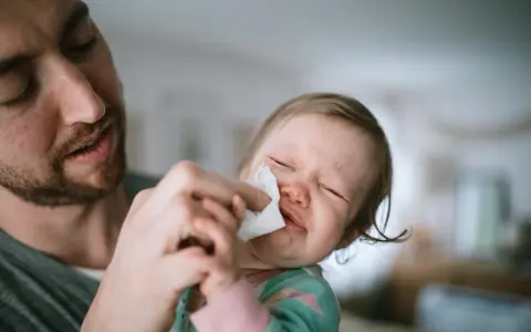 Tata delikatnie wyciera nosek maluszkowi, który ma zielony katar. Dziecko płacze, ale czuje się lepiej.
