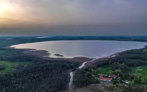 Rozległe jezioro Tuchlin otoczone lasem, z widocznym budynkiem i drogą. Zachód słońca maluje niebo.