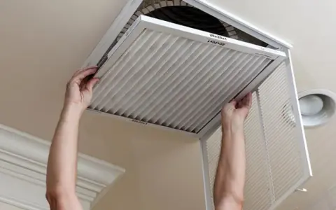 Man hands removing a pleated air filter from a ceiling vent. Essential for limpiar filtros aire acondicionado.