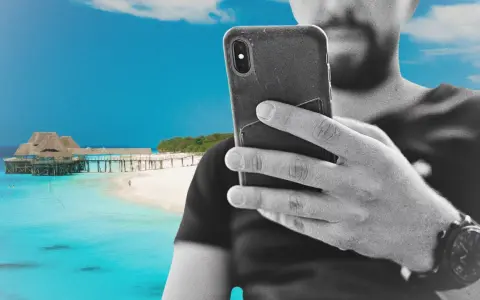 Mężczyzna z telefonem na plaży na Zanzibarze, sprawdzający zanzibar internet. W tle turkusowa woda i drewniany pomost.