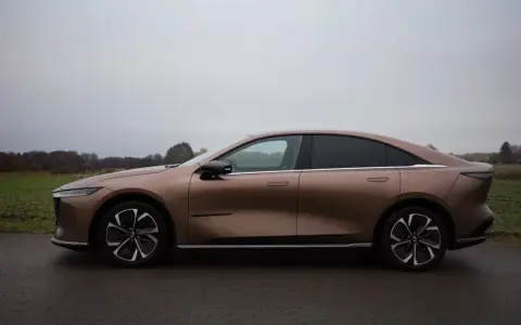 Brązowy samochód elektryczny Mazda6e widziany z boku na tle pochmurnego nieba i krajobrazu.