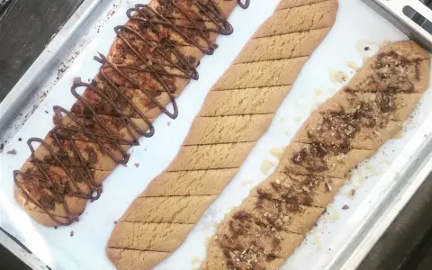 Tre långa, avlånga kakor på en plåt. En är dekorerad med chokladglasyr och hackad choklad, en annan med strössel och en tredje med honung och nötter. Perfekt för ett kolakakor recept!