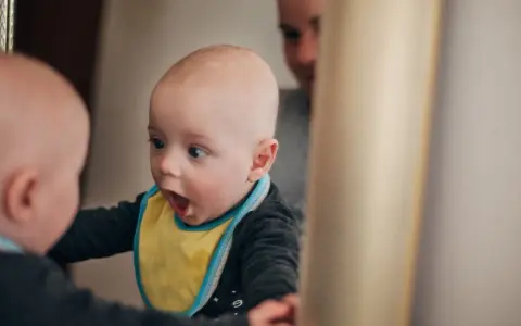 Ein Baby mit gelbem Lätzchen schaut überrascht in einen Spiegel. Vielleicht fragt es sich, ab wann Kinder "Ich" sagen.