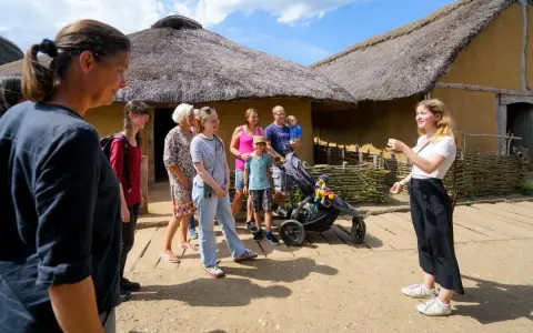 Familienurlaub in Haithabu: Eine Gruppe erkundet historische Häuser mit Strohdächern, während eine junge Frau eine Führung gibt.