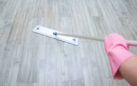 Mano con guante rosa limpiando suelo vinílico con mopa.