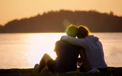 Ein Paar sitzt am Seeufer im Sonnenuntergang, die Arme umeinander geschlungen. Sie finden Trost und lernen, sich selbst zu verzeihen.