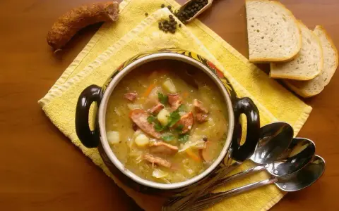 Aromaticzny kapuśniak z kiełbasą i ziemniakami, podany w ozdobnym naczyniu, obok kromek chleba i ziaren pieprzu.