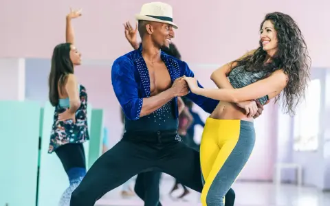 Para tańczy w rytm muzyki, wykonując dynamiczny taniec baciata. W tle widać inne pary ćwiczące.