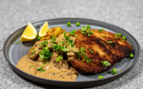 En gyllene panerad kotlett serveras med krämig svampsås, rostade potatisar, ärtor och citronklyftor.