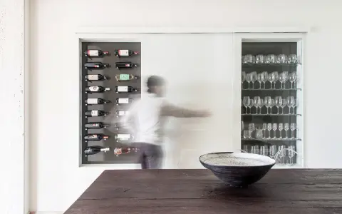 Una nicchia nel muro portante ospita una cantinetta per vini e una vetrina di bicchieri. Una figura sfocata si muove tra i due spazi.