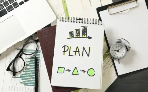 Na biurku leżą dokumenty, laptop, okulary i budzik. Na notatniku widnieje napis "PLAN" i schemat, który symbolizuje indywidualny plan rozwoju.