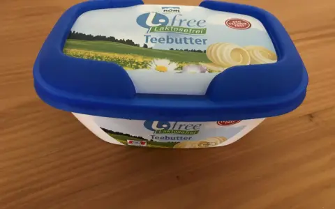 Eine Packung "Biofree Laktosefrei Teebutter" mit blauem Deckel steht auf einem Holztisch. Die Verpackung zeigt eine Sommerwiese mit Blumen und Butterflocken.