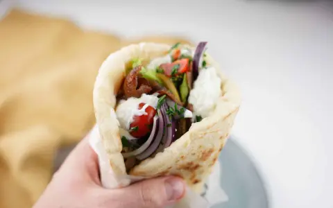 En hand håller en gyros med kött, grönsaker och vit sås. Perfekt för supergod fredagsmat!