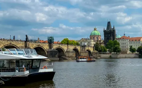 Widok na rzekę w Pradze z łodzią wycieczkową na pierwszym planie i Mostem Karola w tle.