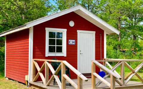 Rött litet hus med vit dörr och fönster, nummer E34. Trädäck med räcke. En grön stol står på däcket. Välkommen till Askeviks Camping!