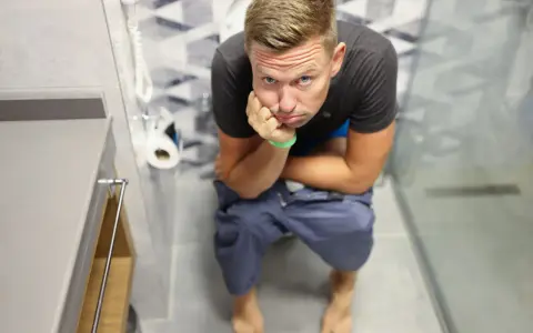 Mężczyzna siedzi na toalecie, podpierając głowę ręką, z miną wyrażającą dyskomfort. Wygląda, jakby czekał na lewatywę przed analem.
