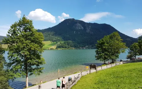 Ein sonniger Tag am bayrischzell see. Menschen spazieren am Ufer entlang, während im Hintergrund ein bewaldeter Berg aufragt.