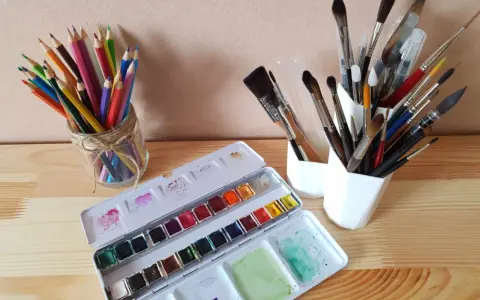Une palette d'aquarelle ouverte, des crayons de couleur dans un bocal et plusieurs pinceaux indispensables aquarelle dans des pots blancs.