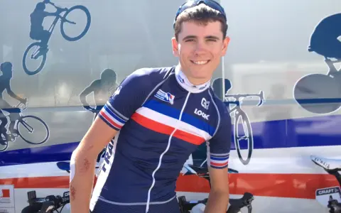 David Gaudu, a French cyclist, smiles in his team's blue, white, and red jersey, with bike graphics on the vehicle behind him.