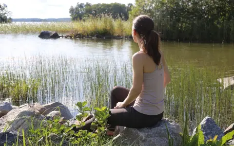 Kvinna sitter vid en sjö, omgiven av vass och stenar. En stund av avslappning mot ångest i naturens lugn.