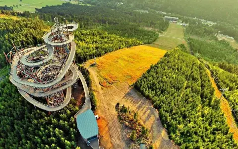 Drewniana ścieżka w koronach drzew w Czechach, przypominająca spiralny sky bridge, otoczona lasem i złotym polem.