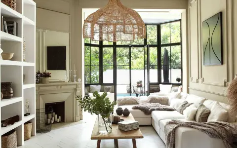 Salon lumineux au style bohème chic maison, avec canapé blanc, suspensions en rotin, cheminée et grandes fenêtres donnant sur le jardin.