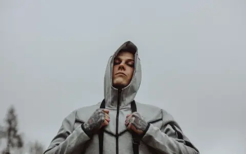 Młody mężczyzna w szarej bluzie z kapturem, z tatuażami na dłoniach, wygląda na zamyślonego. Może to symbol walki z chorobą, jak szpaku choroba, która go trapi.