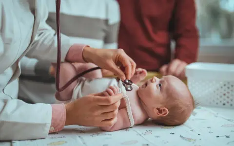 Pielęgniarka środowiskowa po porodzie osłuchuje noworodka. Rodzice obserwują badanie.