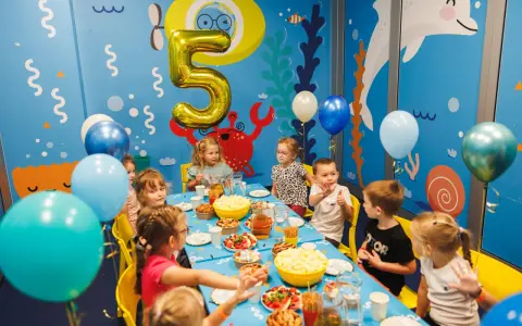 Dzieci świętują 5 urodziny przy stole pełnym przekąsek. Złoty balon w kształcie cyfry 5 unosi się nad stołem.