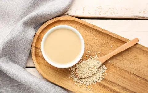 Eine Schale cremiger Tahin neben Sesamsamen auf einem Holzlöffel. So sieht die Antwort auf die Frage "was ist Tahin" aus.