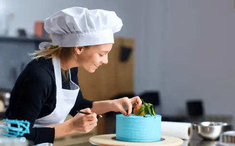 Uśmiechnięta cukierniczka w białym czepku i fartuchu dekoruje błękitny tort. Jej cv to pasja i precyzja w tworzeniu słodkich arcydzieł.