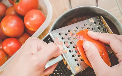 Ręce trą pomidora na tarce, przygotowując go do sosu. Obok stoi miska pełna świeżych pomidorów.