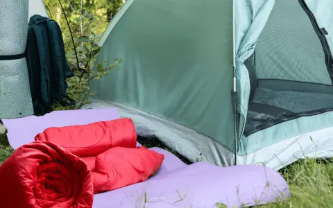 Zwei rote Schlafsäcke liegen auf lila Isomatten vor einem grünen Zelt. Ein Rucksack und eine weitere Isomatte stehen daneben.