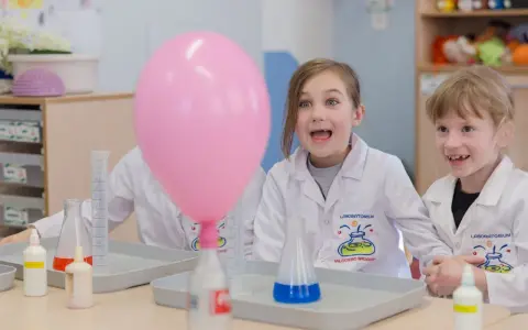 Dzieci w fartuchach laboratoryjnych z ekscytacją obserwują dmuchany balon podczas eksperymentu. To świetna chemia dla dzieci!