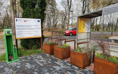 Zielona stacja serwisowa dla rowerów i tablica informacyjna o projekcie VeloMetropolis w Małopolsce. W tle czerwony samochód i jesienne drzewa.