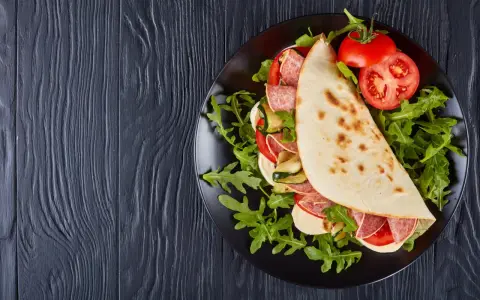 Piadina ripiena con salame, mozzarella, zucchine grigliate e pomodoro, servita su un piatto nero con rucola. Un assaggio della città di Rimini in un ristorante.