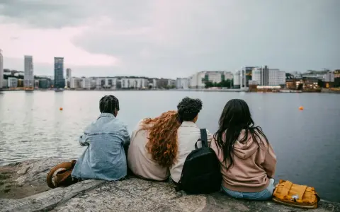 Three young people sit on rocks by the water, looking at a city skyline.
