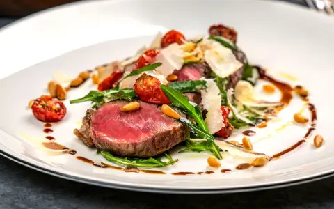 Ein saftiges Steak, italienisch zubereitet, mit Rucola, Kirschtomaten, Pinienkernen und Parmesanspänen auf einem weißen Teller.