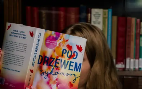 Dziewczyna czyta książkę "Pod drzewem sykomory", ukrywając twarz za okładką. W tle widać regały pełne książek.