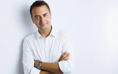 Uśmiechnięty mężczyzna w białej koszuli, z rękami skrzyżowanymi na piersi. Idealne zdjęcie do CV, które buduje zaufanie.