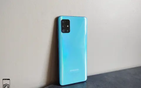 Błękitny Samsung A51 z aparatem w kwadratowej ramce, oparty o jasną ścianę.