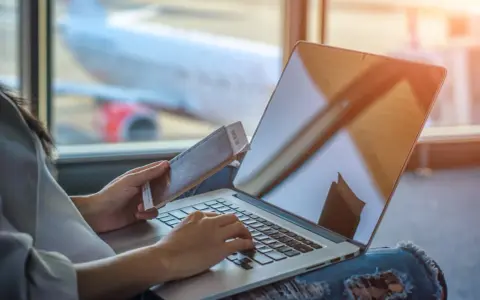 Osoba z paszportem i biletem czeka na lotnisku, sprawdzając na laptopie, ile czasu na odbiór paszportu.
