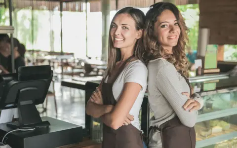Dwie uśmiechnięte pracownice kawiarni, gotowe do obsługi. Od ilu lat można pracować w gastronomii? Odpowiedź znajdziesz w przepisach.
