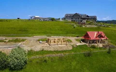 Budowa domu z bali w górach. Widać fundamenty i ściany z bali, a także dom z czerwonym dachem w trakcie prac. To początek budowy, jak wyczyścić dom z bali, by służył latami.